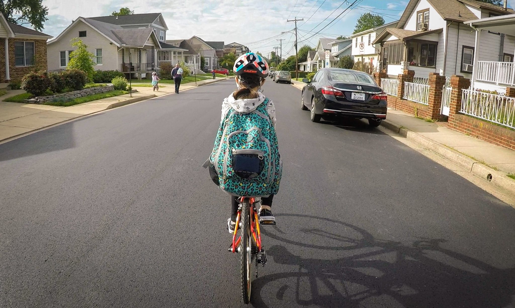 A young girl riding her bike on a calm neighborhood street, wearing a helmet and a large colorful book bag.