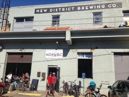 Two men in bike helmets stand together in front of a brewery, converted from an old warehouse.