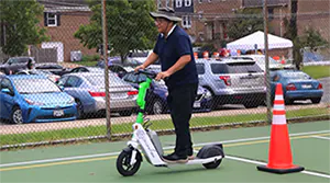 a photo of a guy with a bucket hat riding an e-scooter on a tennis court around an orange street cone