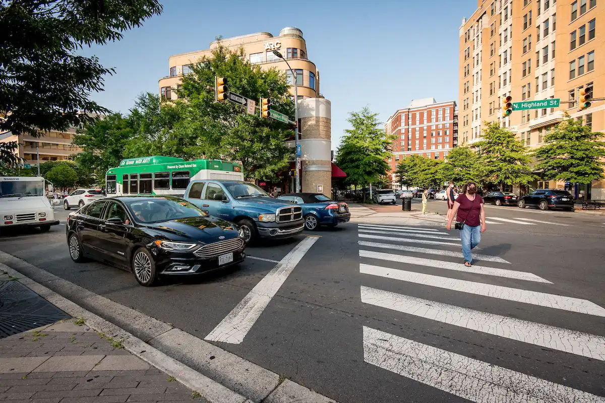 a young lady walking across a crosswalk while cars have all lined up and stopped at a red light on a sunny day